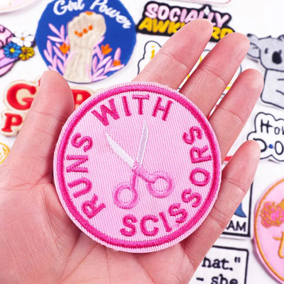 Pink patch with scissors and text held in a hand, surrounded by other patches.
