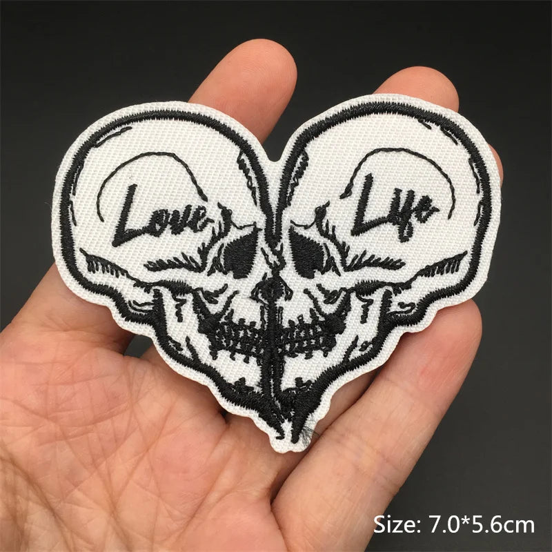 Heart-shaped skull patch with 'Love' and 'Life' text held in a hand against a dark background.