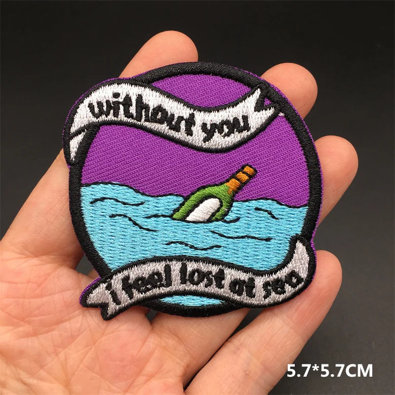 Hand holding a purple blue black and white embroidered Without You I Feel Lost at Sea bottle iron-on patch approximately 5.7x5.7cm on a dark background