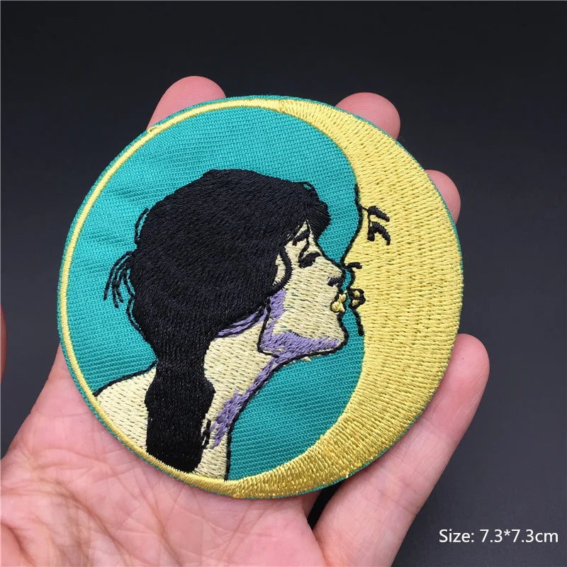 Hand holding a turquoise gold black and lavender embroidered woman kissing crescent moon iron-on patch approximately 7.3x7.3cm on a dark backgroundbackground.