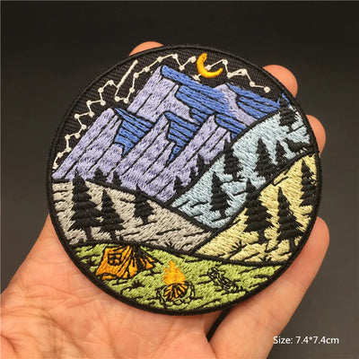 Circular embroidered patch with mountain and nature design held in a hand against a dark background