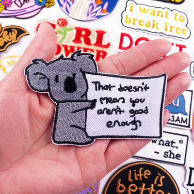 Hand holding a patch with a koala and text, surrounded by other patches.