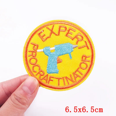 Patch with 'Expert Procrastinator' text and a drill symbol, held by a hand on a white background.