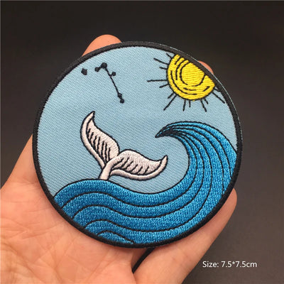 Embroidered patch with whale tail, sun, and waves held in a hand against a dark background
