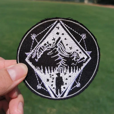 Embroidered patch with a design of a person standing in front of trees held by a hand against a green outdoor background.