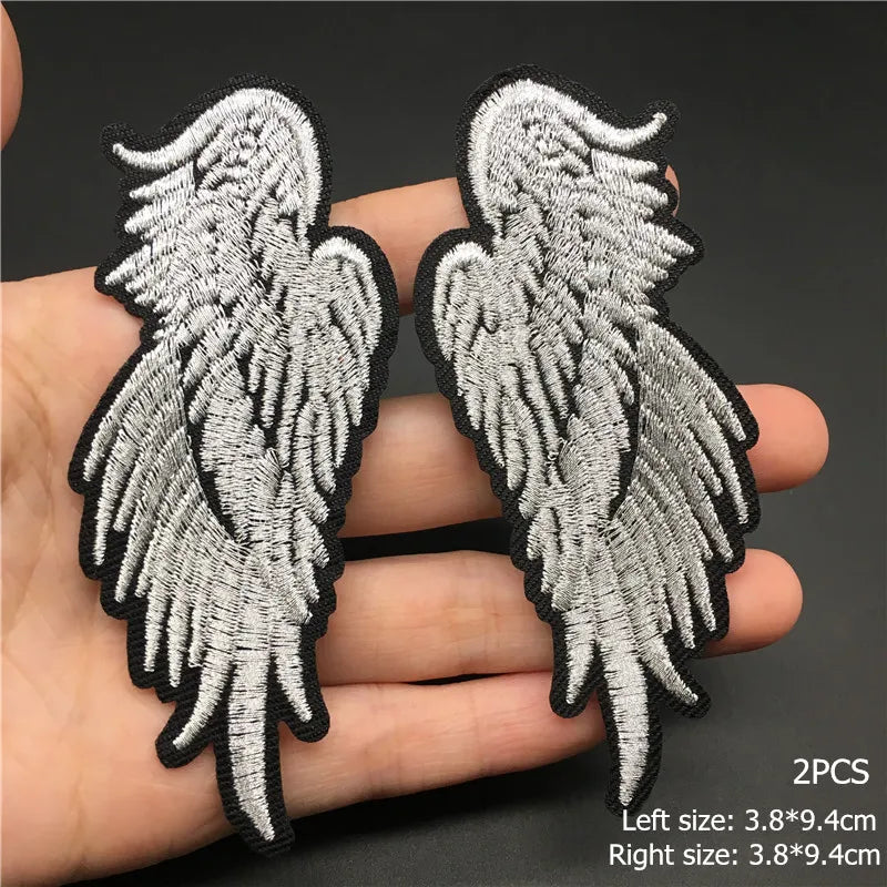 Two silver angel wing patches held between fingers with size information on a dark background.