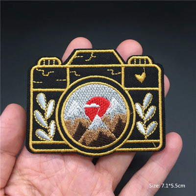 Camera-shaped patch with embroidered design held in a hand against a dark background