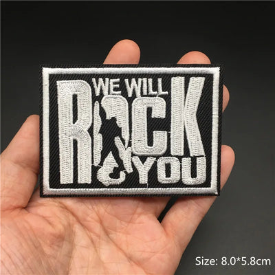 Embroidered patch with 'We Will Rock You' text held in a hand against a dark background