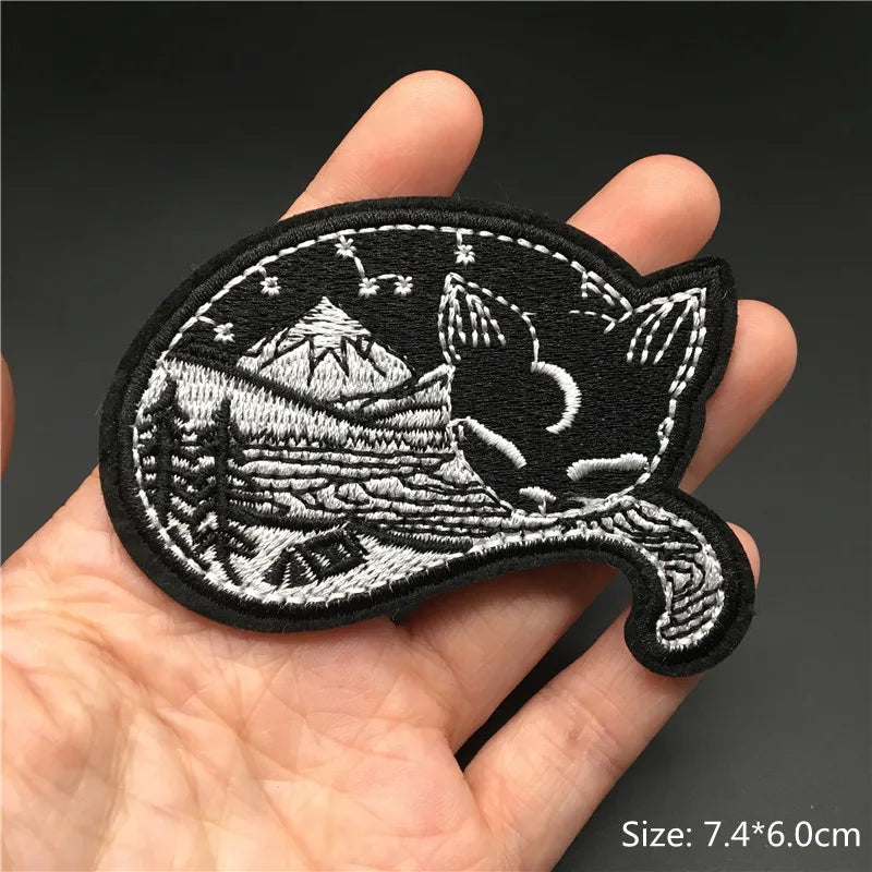 Embroidered patch with mountain and cat design held in a hand against a dark background