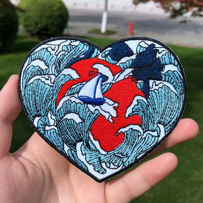 Heart-shaped patch with shark and wave design held in a hand outdoors