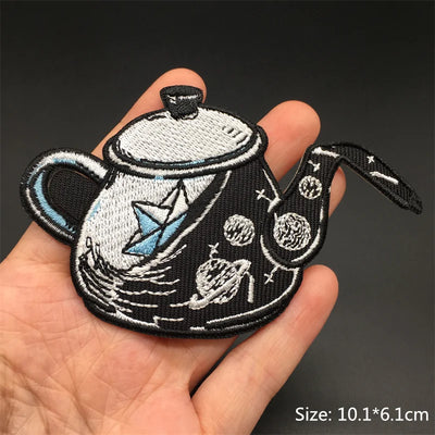 Embroidered patch of a teapot with space-themed design held in a hand against a dark background.
