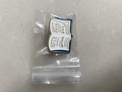 Pin shaped like an open book with text in a clear plastic bag on a gray background