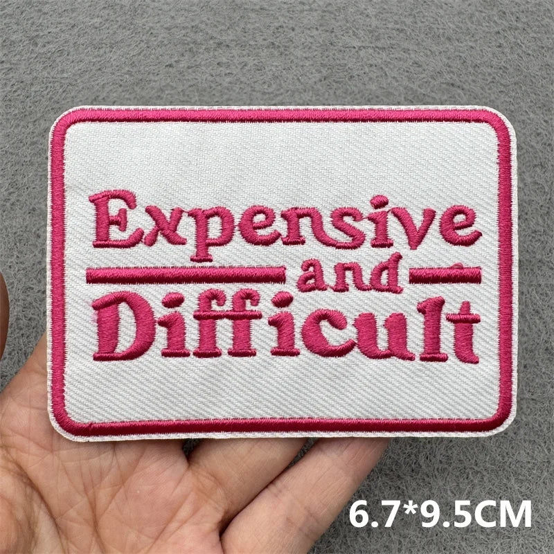 Embroidered patch with 'Expensive and Difficult' text held by a hand on a gray background