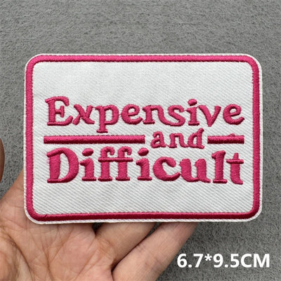 Embroidered patch with 'Expensive and Difficult' text held by a hand on a gray background