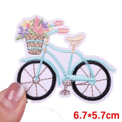 Embroidered patch of a bicycle with a basket of flowers held by a hand on a white background.