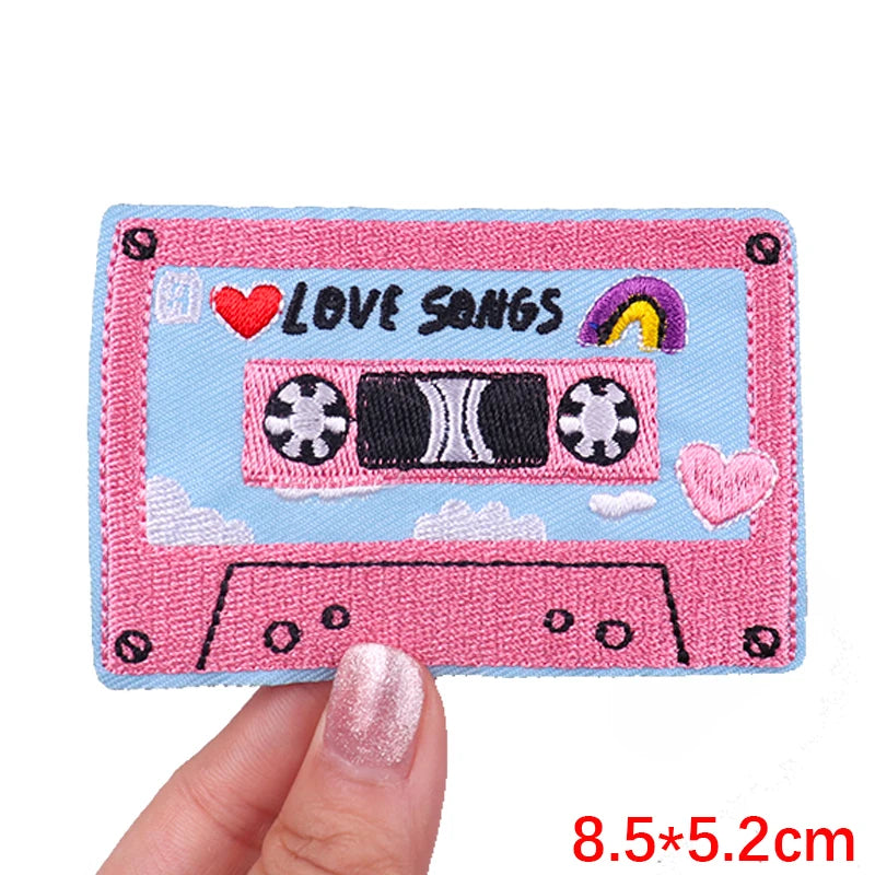 Pink cassette tape patch with 'Love Songs' text held by a hand on a white background