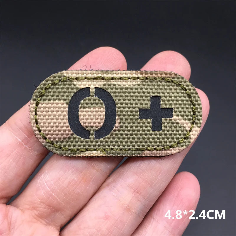 Hand holding an O+ multicam IR reflective tactical blood type hook and loop patch approximately 4.8x2.4cm on a dark background