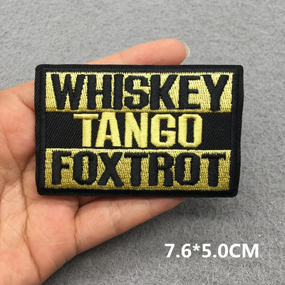 Embroidered patch with 'WHISKEY TANGO FOXTROT' text held in a hand on a gray background.