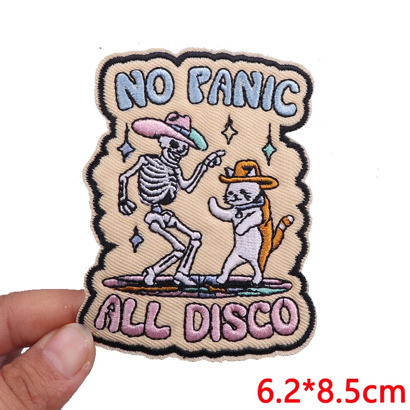 Embroidered patch with skeleton and cat dancing, text 'No Panic All Disco', held by a hand.