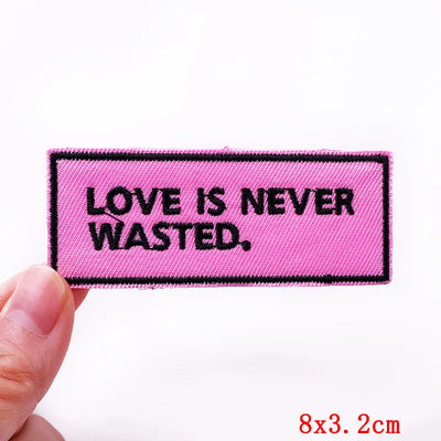 Pink patch with 'LOVE IS NEVER WASTED.' text held by a hand on a white background