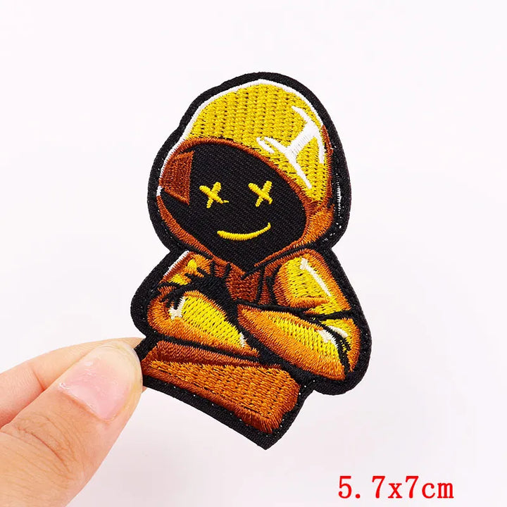 Embroidered patch of a character in a hood with a yellow cap held by a hand on a white background.