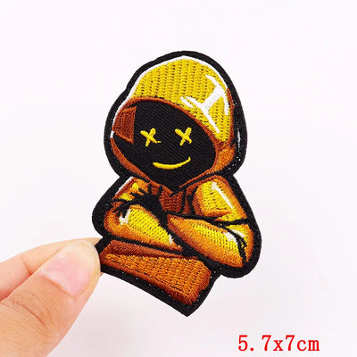 Embroidered patch of a character in a hood with a yellow cap held by a hand on a white background.