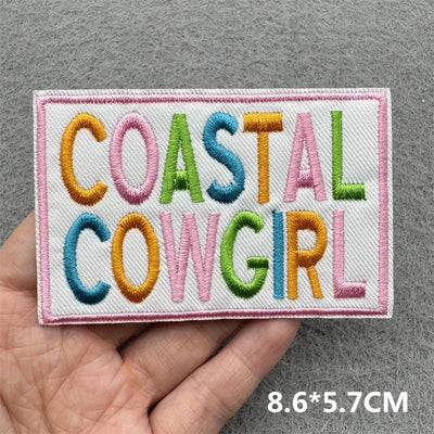 Embroidered patch with 'COASTAL COWGIRL' text held in a hand on a gray background