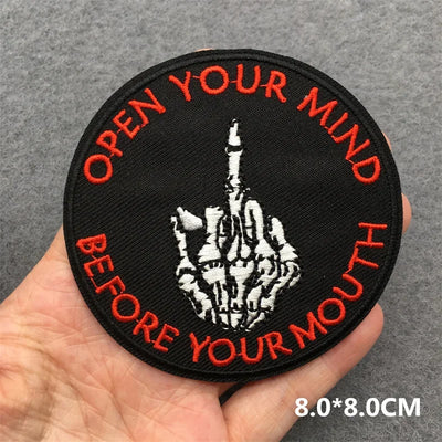 Patch with text 'Open your mind before your mouth' held by a hand on a gray background