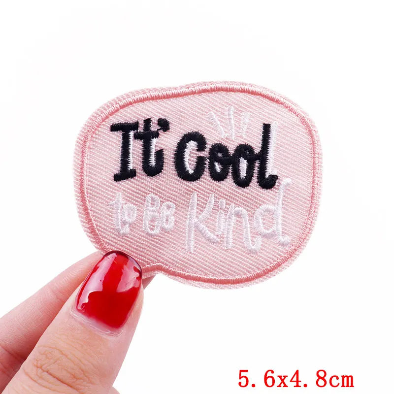 Embroidered patch with 'It's Cool to be Kind' text held by a hand with red nail polish on a white background.