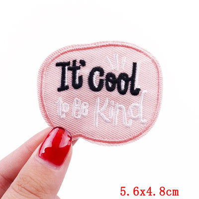 Embroidered patch with 'It's Cool to be Kind' text held by a hand with red nail polish on a white background.