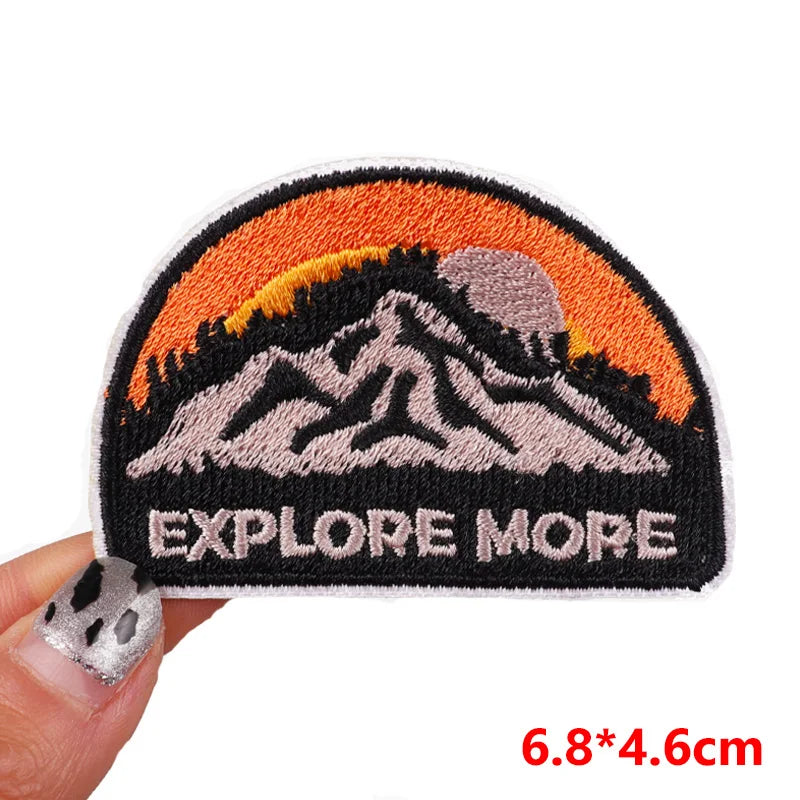 Embroidered patch with mountain design and 'EXPLORE MORE' text held by a hand.