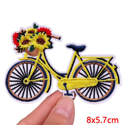 Embroidered patch of a bicycle with flowers in the basket held by a hand on a white background.