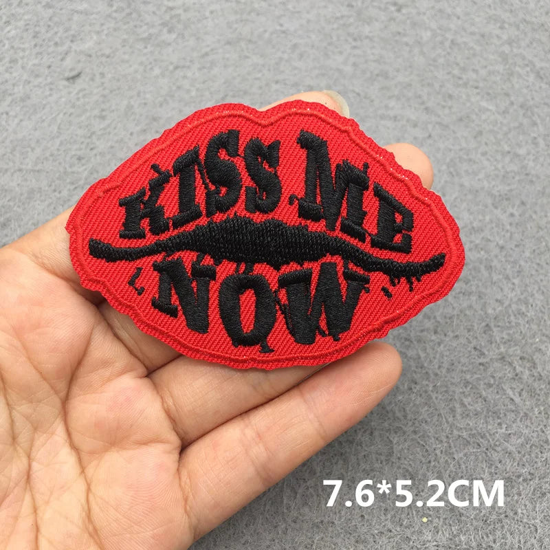 Hand holding a red and black embroidered Kiss Me Now lips iron-on patch approximately 7.6x5.2cm on a gray background