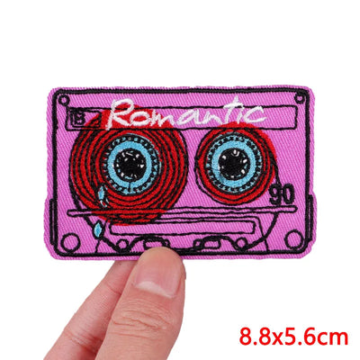 Pink cassette tape patch with 'Romantic' text held by a hand, on a white background.