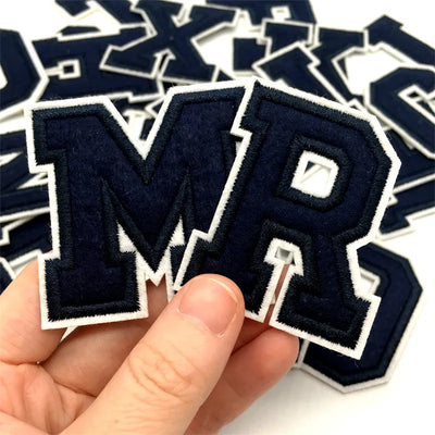 Navy blue and white letter patches with 'MR' held by a hand against a background of similar patches.