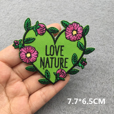 Heart-shaped patch with 'LOVE NATURE' text and flowers held in a hand on a gray background.