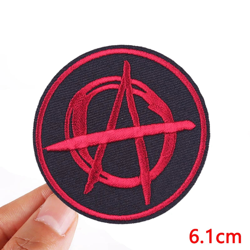 Red anarchy symbol patch on a black background held by a hand, with a measurement label '6.1cm'.