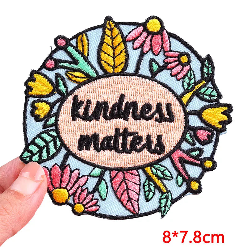Embroidered patch with 'kindness matters' text surrounded by flowers and leaves, held by a hand.