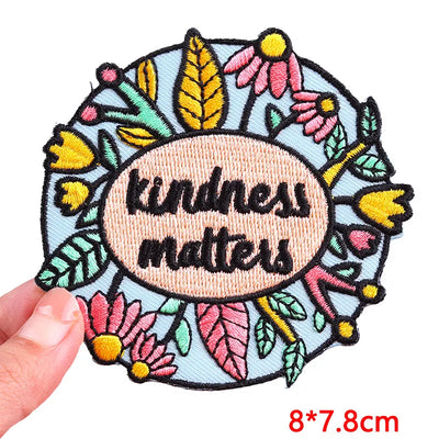 Embroidered patch with 'kindness matters' text surrounded by flowers and leaves, held by a hand.