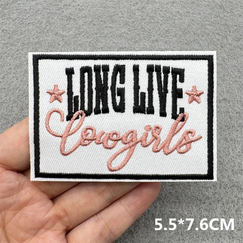 Hand holding a black white and pink embroidered Long Live Cowgirls iron-on patch approximately 5.5x7.6cm on a gray background