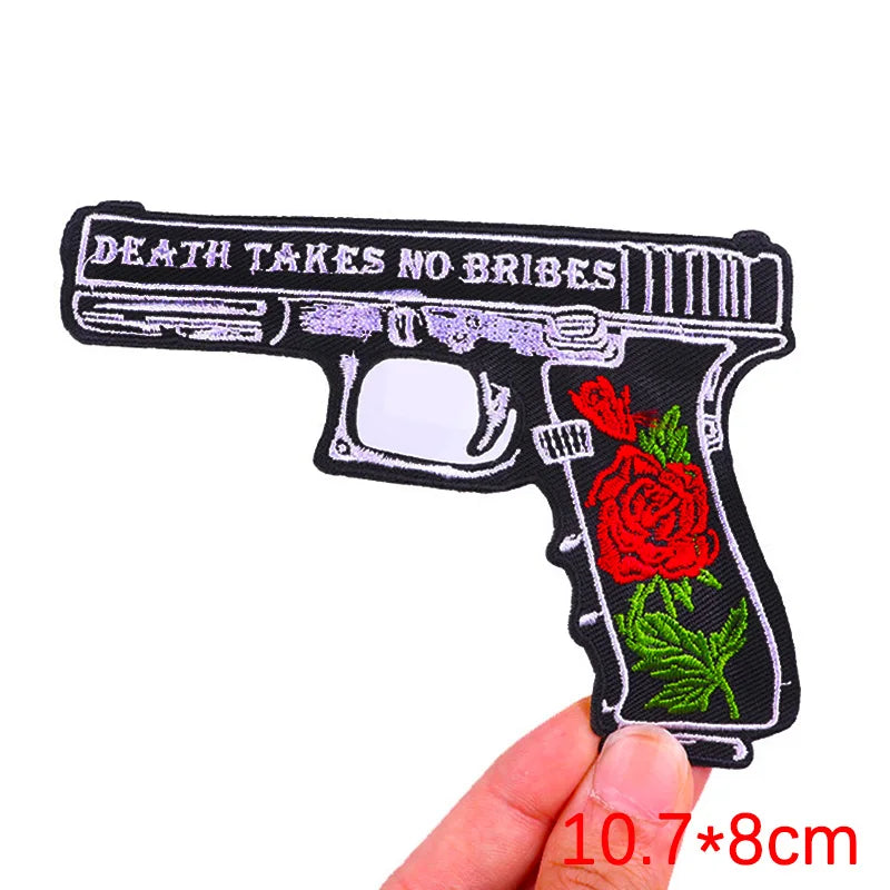 Embroidered patch of a gun with roses and 'Death Takes No Bribes' text, held by a hand.