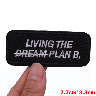 Black patch with 'LIVING THE DREAM PLAN B.' text held by a hand on a white background