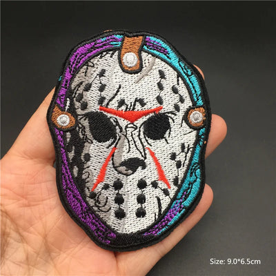 Embroidered patch of a mask with colorful border held in a hand against a dark background