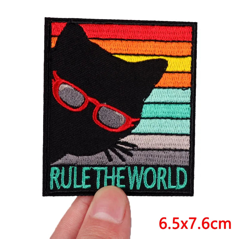 Patch with a black cat wearing red glasses and 'Rule the World' text, held by a hand.