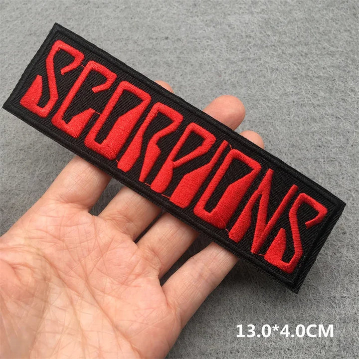 Hand holding a black and red Scorpions embroidered iron-on patch 13x4cm on a grey background