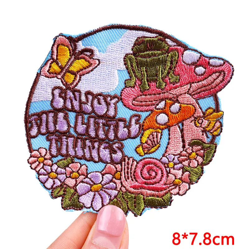 Embroidered patch with mushrooms, flowers, and text held by a hand on a white background
