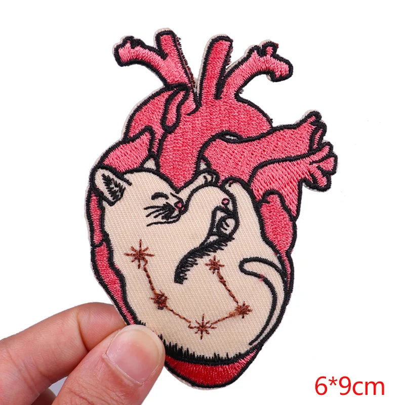 Embroidered patch of a cat inside a heart with a hand holding it, on a white background.