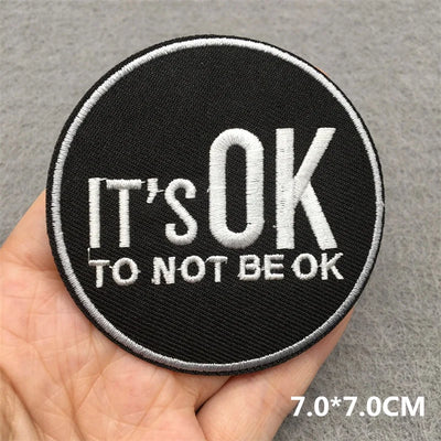 Round black patch with 'IT'S OK TO NOT BE OK' text held in a hand against a gray background.