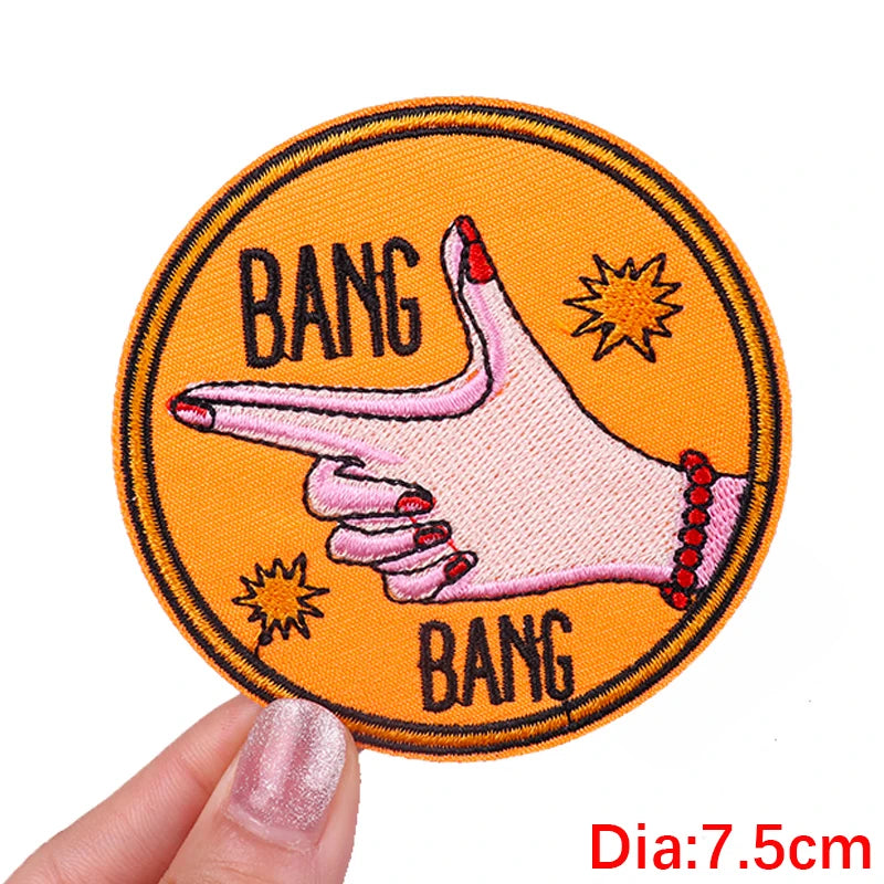 Circular patch with a hand pointing and 'BANG' text on an orange background, held by a hand.