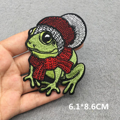 Embroidered patch of a frog wearing a red scarf and hat held by a hand on a gray background.